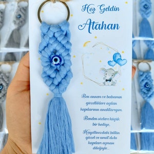 May include: A blue and white crocheted keychain with a blue evil eye charm and a tassel. The keychain is attached to a silver key ring. The keychain is on a white card with the text "Hoş Geldin Atahan" and a message in Turkish.