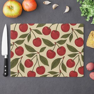 May include: Rectangular cutting board with a repeating pattern of red cherries and green leaves on a beige background. A black-handled knife is on the left side of the board. The board is surrounded by fresh produce.