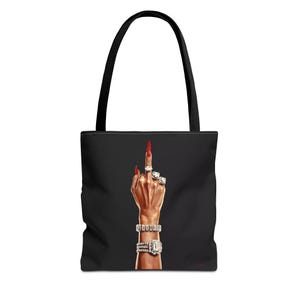 Puede incluir: Bolsa de tela negra con una mano haciendo el dedo medio, con anillos y pulseras de diamantes. La mano tiene esmalte de uñas rojo.