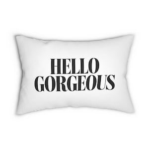 Puede incluir: Cojín rectangular blanco con la frase "HELLO GORGEOUS" en letras negras y en negrita. El cojín tiene un aspecto suave y esponjoso, lo que sugiere una tela cómoda. El diseño es minimalista y contemporáneo, ideal para un hogar moderno.