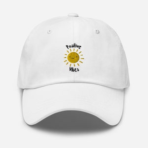 Könnte beinhalten: Weiße Baseballkappe mit einer gestickten gelben Sonne mit einem lächelnden Gesicht und dem Text "Positive Vibes".