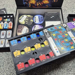 Puede incluir: Juego de mesa con cartas, fichas y un tablero. La caja negra contiene piezas de juego coloridas. El juego parece tener temática de fantasía o aventura. El set incluye cartas con el texto "Creatures from the Black Lagoon".