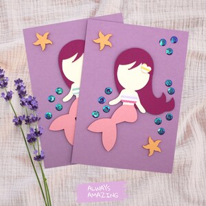 Op de afbeelding: Twee paarse kaarten met elk een uitsparing van een zeemeermin. De zeemeerminnen zijn roze en hebben rood haar. De kaarten zijn versierd met gouden glittersterren en blauwe glitterbellen. De tekst onderaan de kaarten luidt "Altijd geweldig".