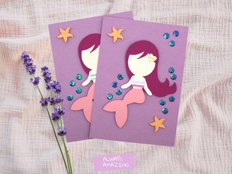 Op de afbeelding: Twee paarse kaarten met elk een uitsparing van een zeemeermin. De zeemeerminnen zijn roze en hebben rood haar. De kaarten zijn versierd met gouden glittersterren en blauwe glitterbellen. De tekst onderaan de kaarten luidt "Altijd geweldig".