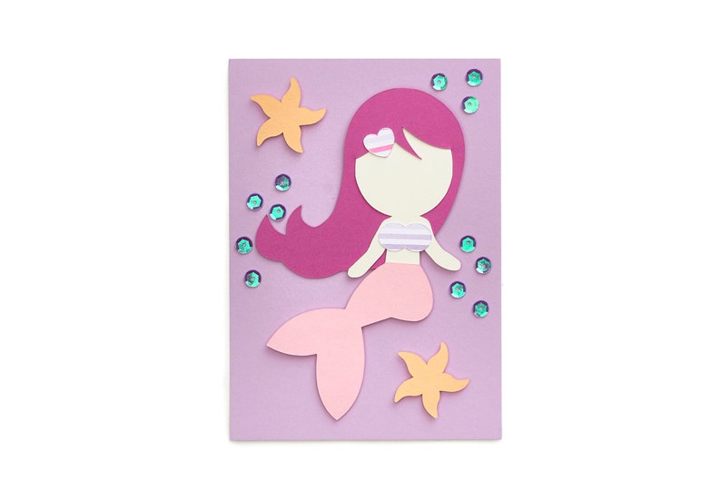 Op de afbeelding: Een paarse kaart met een roze zeemeermin met een roze staart, roze haar en een wit topje met een roze hart op haar hoofd. De zeemeermin wordt omringd door roze en oranje zeesterren en blauwe glitter.