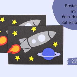 May include: A craft set with two black cards featuring a rocket ship, planets, and stars. The text on the card reads "Basteleset im 6er oder 9er Set erh&auml;ltlich". The text "ALWAYS AMAZING" is on a purple background.
