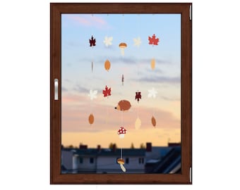 May include: A decorative window mobile featuring autumn-themed cutouts. The mobile includes leaf shapes in red, white, and brown, along with a mushroom and a hedgehog. The mobile hangs in front of a window with a sunset view.