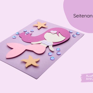 Op de afbeelding: Een paarse kaart met een papieren uitsnijding van een zeemeermin met roze haar, een roze staart en een wit topje. De zeemeermin is omringd door roze en blauwe glitters en oranje zeesterren.