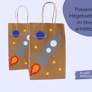 May include: Two brown paper gift bags with handles. Each bag has a rocket ship, planets, and stars cut out of colored paper and glued on. The text "Passende Mitgebselt&uuml;ten im Shop erh&auml;ltlich" is on the right side of the image.
