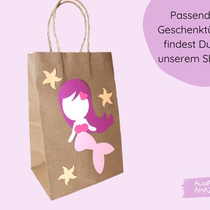 Op de afbeelding: Een bruine papieren cadeauzak met een zeemeerminontwerp. De zeemeermin is gemaakt van roze, paars en wit papier. De tas heeft een wit handvat en is versierd met gele papieren sterren. De tekst "Passende Geschenkt&uuml;ten findest Du in unserem Shop!" is zichtbaar in de rechterbovenhoek van de afbeelding. De tekst "ALWAYS AMAZING" is zichtbaar in de rechterbenedenhoek van de afbeelding.