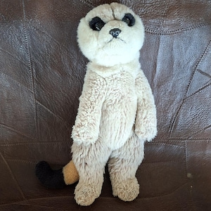 Può includere: Un peluche a forma di suricato con pelliccia color marrone chiaro, macchie marrone scuro intorno agli occhi e un naso nero. L'animale di pezza è in posizione eretta con le braccia lungo i fianchi e una coda con punta nera e base chiara.