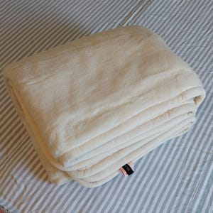 Pode incluir: Uma manta de lã branca dobrada com uma pequena etiqueta preta.