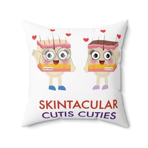 Könnte beinhalten: Ein weißes Kissen mit einem Cartoon-Bild von zwei Hautzellen mit Gesichtern und dem Text "Skintacular Cutis Cuties".