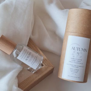 Peut inclure: Une bouteille en verre transparent avec un bouchon en bois et un contenant cylindrique en carton, tous deux étiquetés "AUTUMN". La bouteille est dans un cadre en bois, et le contenant est droit. Le fond est un tissu blanc texturé.