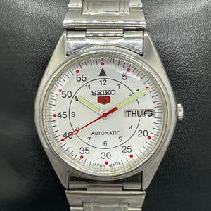 May include: A silver Seiko automatic wristwatch with a white face. The watch has a day-date display, red accents, and a silver strap. The watch face displays the text "SEIKO" and "AUTOMATIC".
