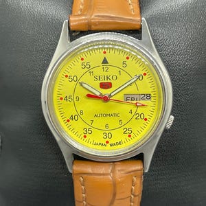 May include: A vintage Seiko automatic wristwatch with a bright yellow face and a brown leather band. The watch has a silver-colored case and features a day and date display. The watch face displays the word "SEIKO" and "JAPAN MADE".