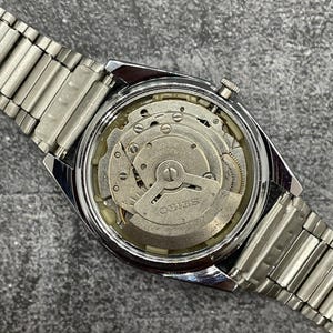 Puede incluir: Un reloj Seiko vintage con una correa de metal plateado. La parte trasera del reloj est&aacute; abierta, revelando el intrincado movimiento mec&aacute;nico con la marca "Seiko" visible. El reloj est&aacute; sobre un fondo gris.