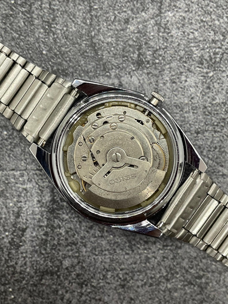 Puede incluir: Un reloj Seiko vintage con una correa de metal plateado. La parte trasera del reloj est&aacute; abierta, revelando el intrincado movimiento mec&aacute;nico con la marca "Seiko" visible. El reloj est&aacute; sobre un fondo gris.