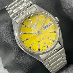 Puede incluir: Un reloj de pulsera autom&aacute;tico Seiko 5 plateado con esfera amarilla. La esfera del reloj muestra el d&iacute;a y la fecha, con el d&iacute;a que indica "JUE 4". El reloj tiene una correa plateada y las palabras "AUTOMATIC" y "JAPAN MADE" son visibles.