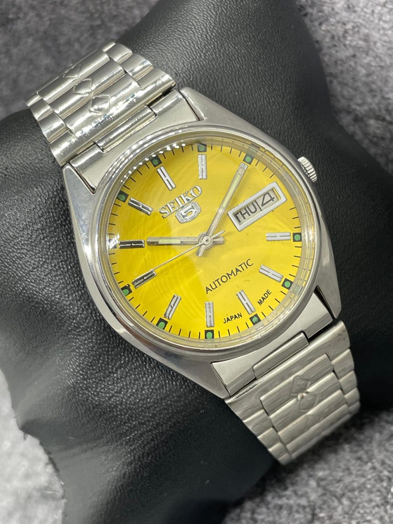 Puede incluir: Un reloj de pulsera autom&aacute;tico Seiko 5 plateado con esfera amarilla. La esfera del reloj muestra el d&iacute;a y la fecha, con el d&iacute;a que indica "JUE 4". El reloj tiene una correa plateada y las palabras "AUTOMATIC" y "JAPAN MADE" son visibles.