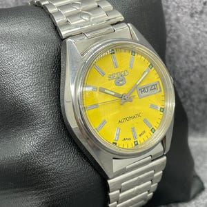 Puede incluir: Reloj de pulsera autom&aacute;tico Seiko plateado con esfera amarilla brillante. El reloj presenta una pantalla de d&iacute;a y fecha, una correa plateada y el texto "SEIKO" y "AUTOMATIC". El reloj se muestra sobre una superficie negra.
