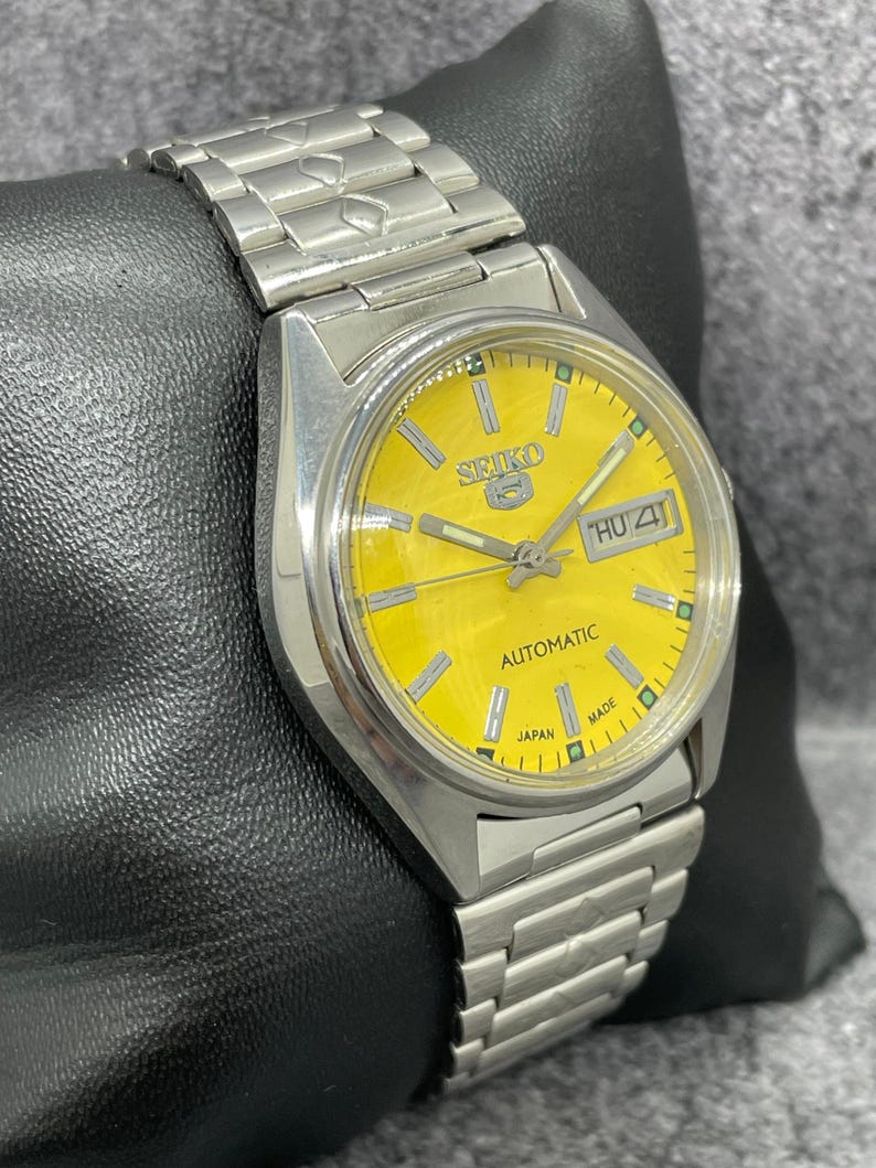 Puede incluir: Reloj de pulsera autom&aacute;tico Seiko plateado con esfera amarilla brillante. El reloj presenta una pantalla de d&iacute;a y fecha, una correa plateada y el texto "SEIKO" y "AUTOMATIC". El reloj se muestra sobre una superficie negra.