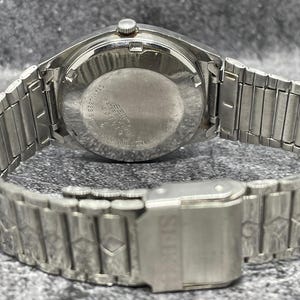 Puede incluir: Un reloj de pulsera plateado con una correa de metal. La esfera del reloj es redonda y la correa est&aacute; hecha de eslabones de metal entrelazados. El cierre es visible y el reloj parece ser vintage.