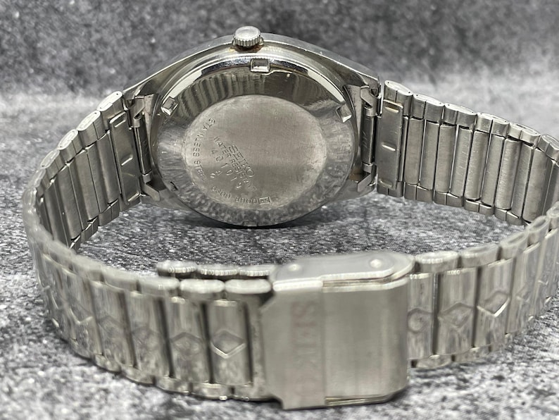 Puede incluir: Un reloj de pulsera plateado con una correa de metal. La esfera del reloj es redonda y la correa est&aacute; hecha de eslabones de metal entrelazados. El cierre es visible y el reloj parece ser vintage.