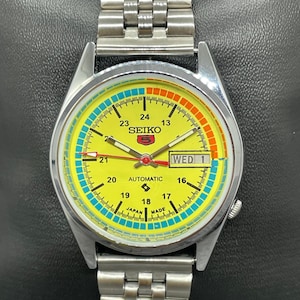 Pode incluir: Um relógio automático Seiko 5 amarelo, prateado e vermelho com exibição do dia e da data. O mostrador do relógio tem um fundo amarelo com um anel exterior azul e vermelho. O relógio tem uma pulseira de metal prateada.