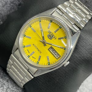 Puede incluir: Un reloj de pulsera autom&aacute;tico Seiko plateado con esfera amarilla brillante. El reloj muestra el d&iacute;a y la fecha, con "THU 4". La correa es de eslabones met&aacute;licos plateados. La esfera tiene el texto "SEIKO" y "JAPAN MADE".