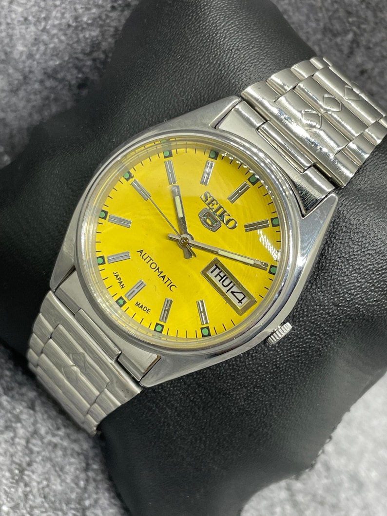 Puede incluir: Un reloj de pulsera autom&aacute;tico Seiko plateado con esfera amarilla brillante. El reloj muestra el d&iacute;a y la fecha, con "THU 4". La correa es de eslabones met&aacute;licos plateados. La esfera tiene el texto "SEIKO" y "JAPAN MADE".