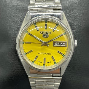 Puede incluir: Un reloj de pulsera autom&aacute;tico Seiko 5 vintage con una esfera de color amarillo brillante. El reloj tiene una caja y una correa de acero inoxidable plateado. La esfera muestra el d&iacute;a y la fecha, con el texto "SEIKO" y "AUTOMATIC".