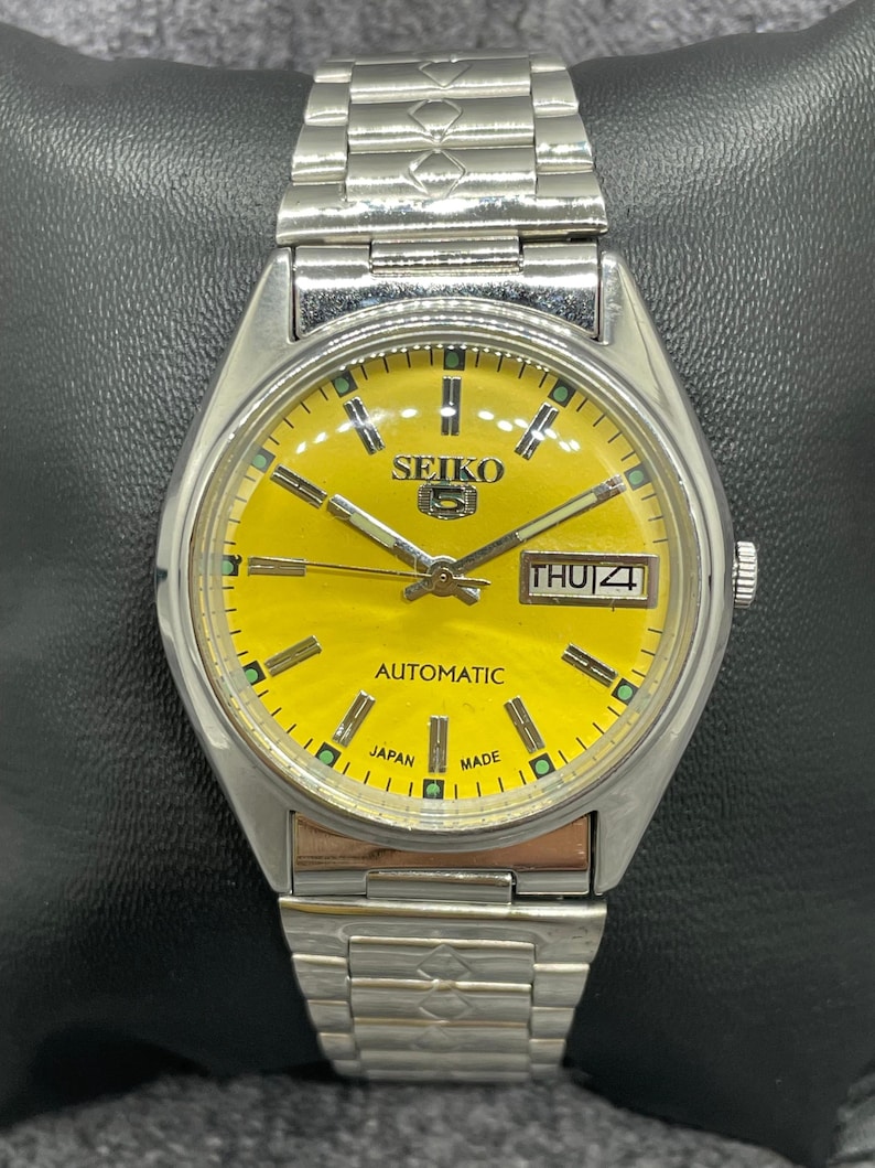 Puede incluir: Un reloj de pulsera autom&aacute;tico Seiko 5 vintage con una esfera de color amarillo brillante. El reloj tiene una caja y una correa de acero inoxidable plateado. La esfera muestra el d&iacute;a y la fecha, con el texto "SEIKO" y "AUTOMATIC".