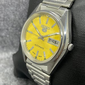 Puede incluir: Un reloj de pulsera autom&aacute;tico Seiko 5 plateado con una esfera amarilla brillante. El reloj presenta una pantalla de d&iacute;a y fecha, manecillas plateadas y un brazalete de eslabones plateados a juego. La esfera indica "Automatic" y "Japan Made".