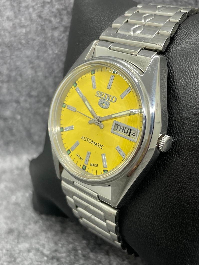 Puede incluir: Un reloj de pulsera autom&aacute;tico Seiko 5 plateado con una esfera amarilla brillante. El reloj presenta una pantalla de d&iacute;a y fecha, manecillas plateadas y un brazalete de eslabones plateados a juego. La esfera indica "Automatic" y "Japan Made".