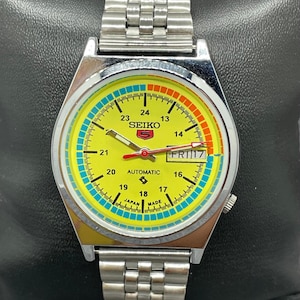 May include: A yellow, silver and red Seiko 5 automatic wristwatch with a day and date display. The watch face has a black and white inner dial with a red second hand. The watch band is a silver metal bracelet.