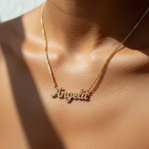 May include: A gold-colored necklace with the name "Angela" spelled out in sparkling, clear gemstones. The pendant hangs from a delicate chain, resting on a person's skin. The necklace is a personalized jewelry item.