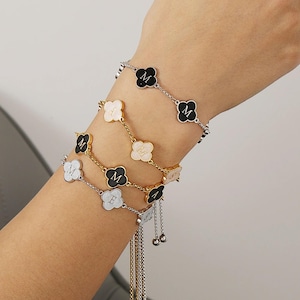 May include: Four gold bracelets with a clover design. Each bracelet has a different colored clover charm: black, white, gold, and a combination of black and white. The charms have the letter 'M' engraved on them.