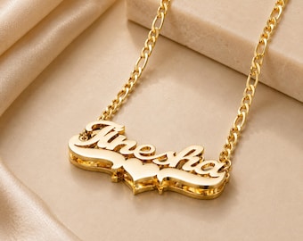 May include: A gold-toned nameplate necklace featuring the name Tinesha in a cursive script font. The pendant includes a small heart shape integrated into the design and is attached to a classic Figaro chain with flat, elongated links.