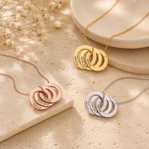 May include: Three necklaces with interlocking circular pendants in gold, rose gold, and silver tones. Each pendant features three rings engraved with the names Marco and Antonia and the date 03.04.2018. The necklaces have delicate matching chains.