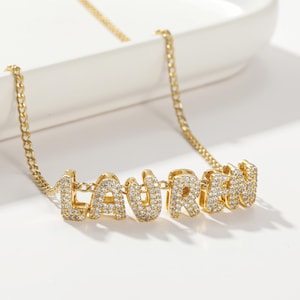 May include: Gold chain necklace with the name "LAUREN" spelled out in large, block letters. Each letter is encrusted with small, clear cubic zirconia stones.