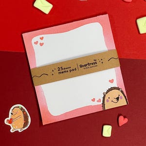 May include: A pink memo pad with a watercolour effect, featuring a smiling hedgehog illustration and small hearts. The pad is held together with a brown paper band that reads "25 sheets memo pad". Decorative elements include heart and envelope-shaped sweets.