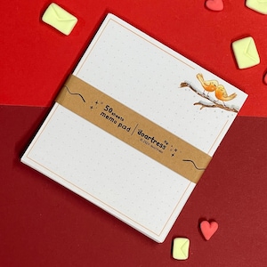 Peut inclure: Un bloc-notes avec un motif à points et une bande marron avec l'inscription "50 feuilles memo pad doartress". Le bloc-notes présente une illustration à l'aquarelle de deux oiseaux jaunes sur une branche. Des décorations en forme de cœur et d'enveloppe sont éparpillées sur un fond rouge.