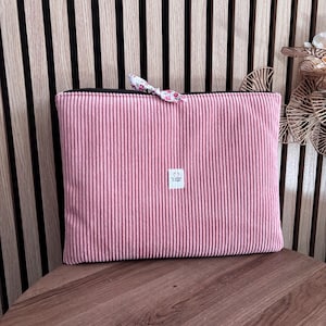 Peut inclure: Une pochette rectangulaire en velours côtelé rose dotée d'une fermeture éclair noire avec une petite tirette en tissu floral. Une étiquette rectangulaire blanche est centrée sur le devant avec le texte La couture de Sophie imprimé en noir.