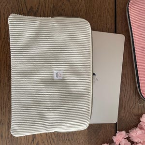 Peut inclure: Une housse d'ordinateur portable en velours côtelé vert clair avec une fermeture éclair. Un ordinateur portable argenté est partiellement inséré. Une petite étiquette est cousue sur le devant. Une housse rose est visible à droite.