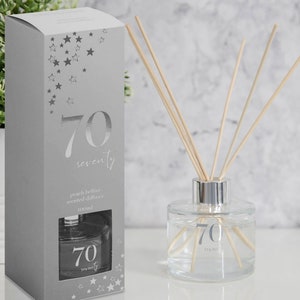 May include: A silver gift box with a silver star pattern and the number 70 printed on it. The box contains a clear glass bottle with a silver lid and a silver label that says "70 seventy" and "peach bellini scented diffuser 100ml". The bottle is filled with a clear liquid and has wooden sticks in it.
