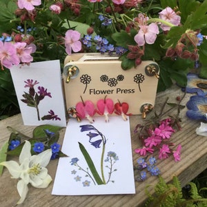 Può includere: Una pressa per fiori in legno con un'etichetta bianca che dice "Flower Press". La pressa è aperta e contiene diversi fiori pressati, tra cui fiori rosa, viola e blu.