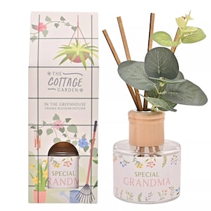 Puede incluir: Un difusor de vidrio con tapa de madera y varillas, etiquetado como "Special Grandma". La botella está decorada con motivos florales y contiene una planta verde. El embalaje dice "The Cottage Garden" y "Orange Blossom Diffuser".