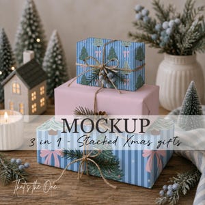 Könnte beinhalten: Drei gestapelte rechteckige Geschenkboxen, eingewickelt in blau gestreiftes Papier mit grünen Tannenbaum- und rosa Schleifenmustern sowie einfarbigem hellrosa Papier. Die Geschenke sind mit Naturkordel, kleinen blauen Beeren und Tannenzweigen verziert.