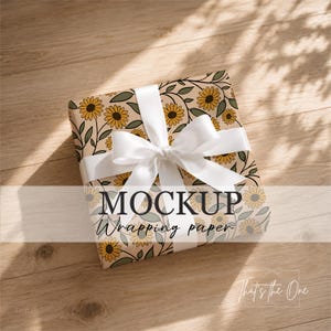 Peut inclure: Une boîte cadeau carrée enveloppée dans du papier beige clair avec un motif répétitif de tournesols jaunes aux centres brun foncé et des feuilles vert sauge. La boîte est nouée avec un ruban de satin blanc. Le texte MOCKUP Wrapping paper est visible.